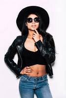 Beauty in style. Beautiful young mixed race woman in leather jacket and hat standing against white background and holding finger on chin photo