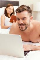 Spending leisure time in bed. Handsome young and shirtless man lying in bed and working on laptop while woman reading a book in the background photo