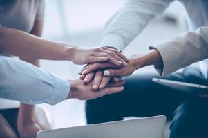 Together we can do everything Close-up part of top view of group of four young people holding hands and showing their unity while sitting at office photo