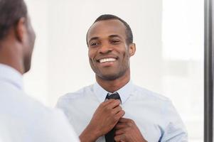 Confident about his look. Young African man adjusting his necktie while standing against mirror photo