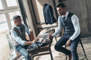 Listening to client requirements. Young fashionable designer choosing fabric with his client while sitting in the workshop photo