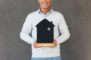 New house owner. Cropped image of mature man holding house shaped object in hand and smiling while standing against grey background photo