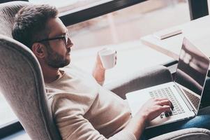 Surfing web from cafe. Side view of handsome man using his laptop and holding coffee cup while sitting in chair in front of window photo