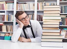 libros para leer. un joven alegre con camisa y corbata de moño sentado en la mesa de la biblioteca y mirando la pila de libros foto