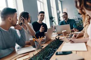 Achieving success together. Group of young modern people in smart casual wear discussing something and smiling while working in the creative office photo