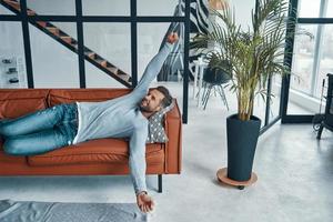 Handsome young man in casual clothing stretching and smiling while lying on the sofa at home photo