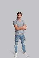 Confidence and charisma. Full length of handsome young man keeping arms crossed and looking at camera while standing against grey background photo