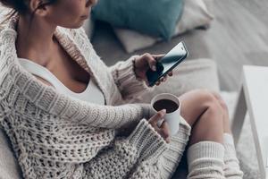 Texting to boyfriend. Close-up of beautiful young woman holding cup and using smart phone while relaxing on couch at home photo