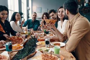 vacaciones entre amigos. grupo de jóvenes con ropa informal comiendo pizza y sonriendo mientras cenan en el interior foto