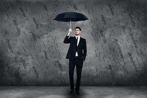 Feeling protected. Full length of confident young man in full suit holding umbrells while standing in front of the concrete wall photo