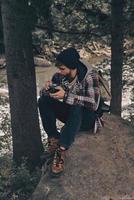 He is opened to adventures. Young modern man with backpack holding a photo camera while sitting in the woods with river in background