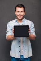 This can help you Cheerful young man shoving his digital tablet with copy space and looking at camera with smile while standing against grey background photo