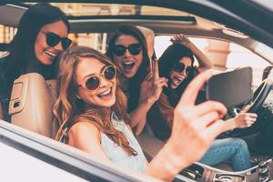 Vista lateral del tiempo de selfie de cuatro hermosas mujeres jóvenes y alegres haciendo selfie y sonriendo mientras están sentadas juntas en el auto foto