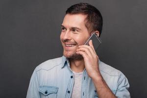 Nice to hear you Cheerful young man talking on mobile phone and looking away with smile while standing against grey background photo