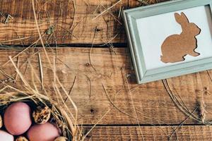 Ester bunny hunt begins Top view of colored Easter eggs in bowl with hay and Easter bunny in picture frame lying on wooden rustic table photo