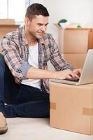 Surfing web in a new house. Handsome young man sitting on the floor and typing something on laptop while cardboard boxes laying on the background photo
