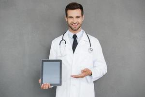 Copy space on his tablet. Cheerful young doctor in white uniform holding digital tablet and pointing at it while standing against grey background photo