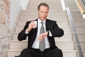 Late again. Worried mature man in formalwear holding a coffee cup and checking a time while sitting on staircase photo