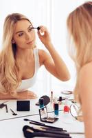 Creating long lashes. Beautiful young woman applying mascara and looking at her reflection in mirror while sitting at the dressing table photo