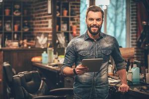 mantener el negocio en la cima con tecnologías digitales. alegre joven barbudo mirando a la cámara y sosteniendo una tableta digital mientras está de pie en la barbería foto