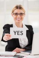 Saying yes to you. Cheerful mature woman in formalwear stretching out paper with yes on it while sitting at her working place photo