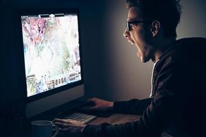 la victoria está cerca. vista lateral de un joven jugando videojuegos mientras está sentado en la mesa en una habitación oscura foto