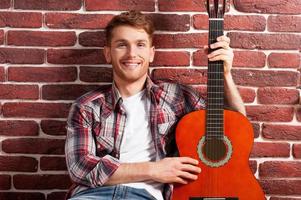 I love my guitar. Handsome young man holding acoustic guitar and smiling while leaning at the brick wall photo
