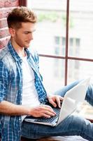 Man surfing the net. Top view of handsome young man working on laptop while sitting on the window sill photo