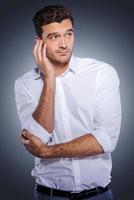 Feeling uncertain. Handsome young man in white shirt holding hand in hair and looking away while standing against grey background photo