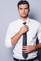 Handsome and self-confident. Handsome young man in formalwear adjusting his necktie and looking at camera while standing against grey background photo