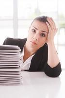 Depressed and overworked. Depressed young woman in suit looking at camera and holding head in hand while sitting at the table with stack of documents laying on it photo