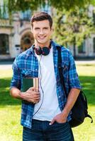 listo para estudiar apuesto joven sosteniendo libros y sonriendo mientras está de pie frente a su universidad foto