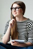 Just inspired. Thoughtful young woman in striped clothing holding note pad and touching her chin with pen while sitting on the hardwood floor photo