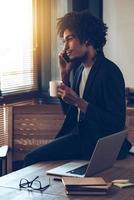 I have some great news for you Side view of young handsome African man talking on mobile phone and holding coffee cup while sitting on table at his working place photo