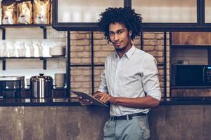 amando su pequeña empresa. joven africano alegre que usa una tableta digital y mira la cámara con una sonrisa mientras está de pie en el mostrador del bar foto