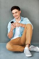 Sending message to friend. Joyful young man holding mobile phone and smiling while sitting against grey background photo