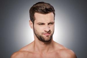 Feeling disgust. Portrait of frustrated young shirtless man looking away and grimacing while standing against grey background photo