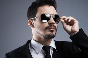 Stylish handsome. Handsome young man in formalwear adjusting his sunglasses and looking away while standing against grey background photo