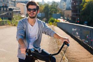 listo para montar. apuesto joven en bicicleta mirando a la cámara y sonriendo mientras está de pie al aire libre foto