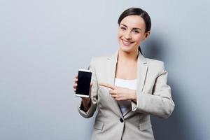 Your advertising on her phone. Beautiful young businesswoman holding mobile phone and pointing on it while standing against grey background photo