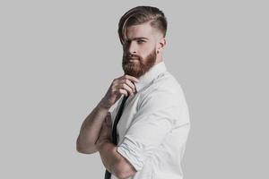 Young and successful. Young businessman keeping hand on chin and looking at camera while standing against grey background photo