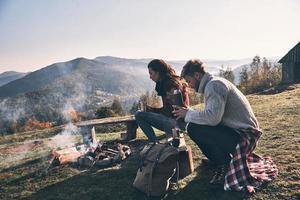 descansando juntos. hermosa pareja joven tomando café por la mañana mientras se sienta junto a la fogata en las montañas foto
