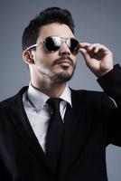 Confident in his style. Handsome young man in formalwear adjusting his sunglasses and looking away while standing against grey background photo