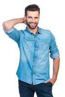 He got candid smile. Confident young handsome man in jeans shirt holding hand behind head and smiling while standing against white background photo