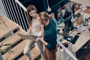 Top view of two young modern colleagues in smart casual wear communicating and using laptop while standing on the stairs photo