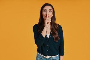 Beautiful young woman in casual holding finger on lips while standing against yellow background photo