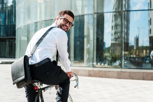 Green way to get to office. Rear view of joyful young businessman looking at camera and smiling while riding on his bicycle with office building in the background photo