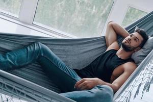 Simply resting. Top view of handsome young man in casual clothing relaxing while lying in the hammock indoors photo