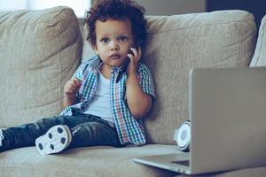 I am so busy Little African baby boy talking on mobile phone and looking at camera while sitting on the couch at home photo