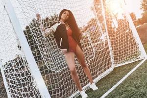 chica de fútbol longitud completa de una joven atractiva con ropa deportiva mirando a la cámara mientras está de pie cerca del poste de la portería foto
